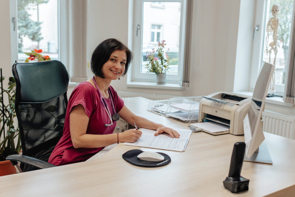 Paní doktorka v červené blůze s fonendoskopem kolem krku vyplňuje papírování na stole v jasně osvětlené lékařské kanceláři.