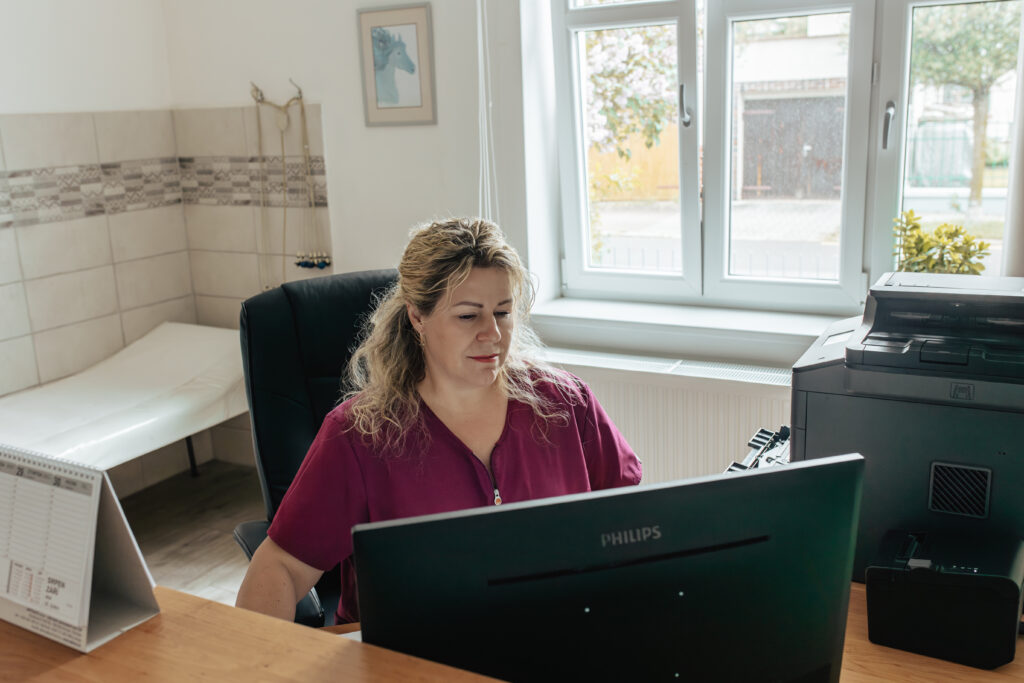 Osoba v růžové košili pracuje na počítači Philips ve světlé a organizované domácí kanceláři s okenním výhledem, vedle stojí tiskárna a na zdi visí umělecké dílo.